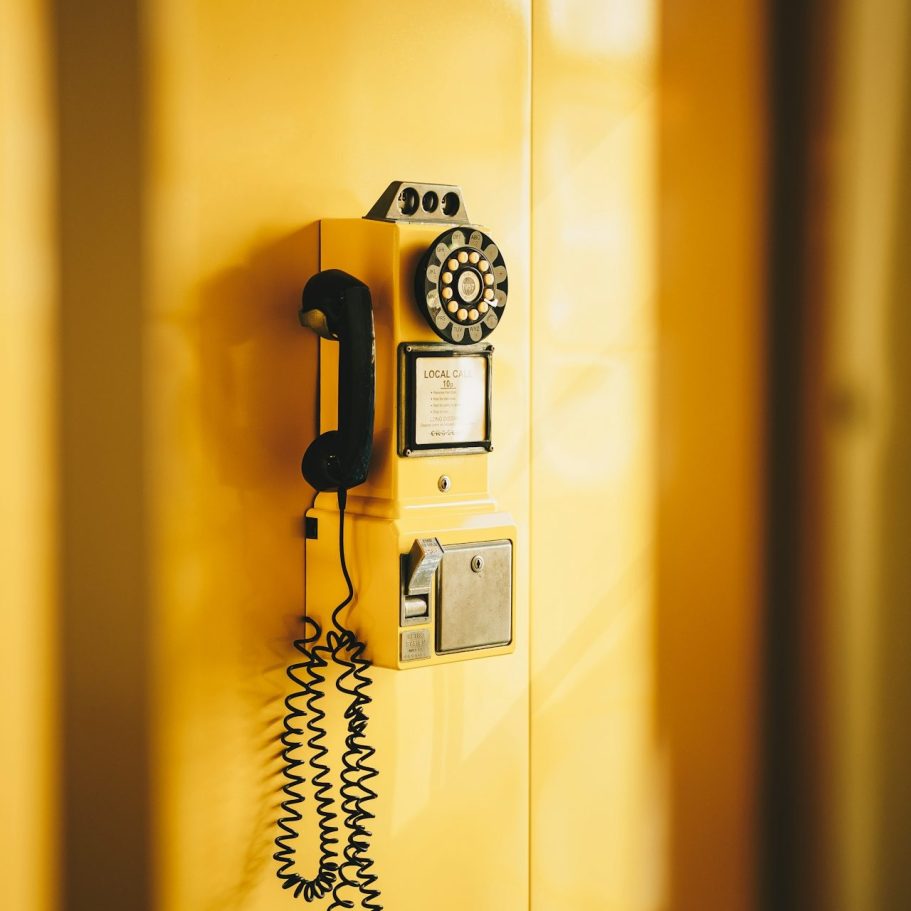 Gelbes Wählscheibentelefon an einer Wand, mit einem Spiralkabel verbunden.